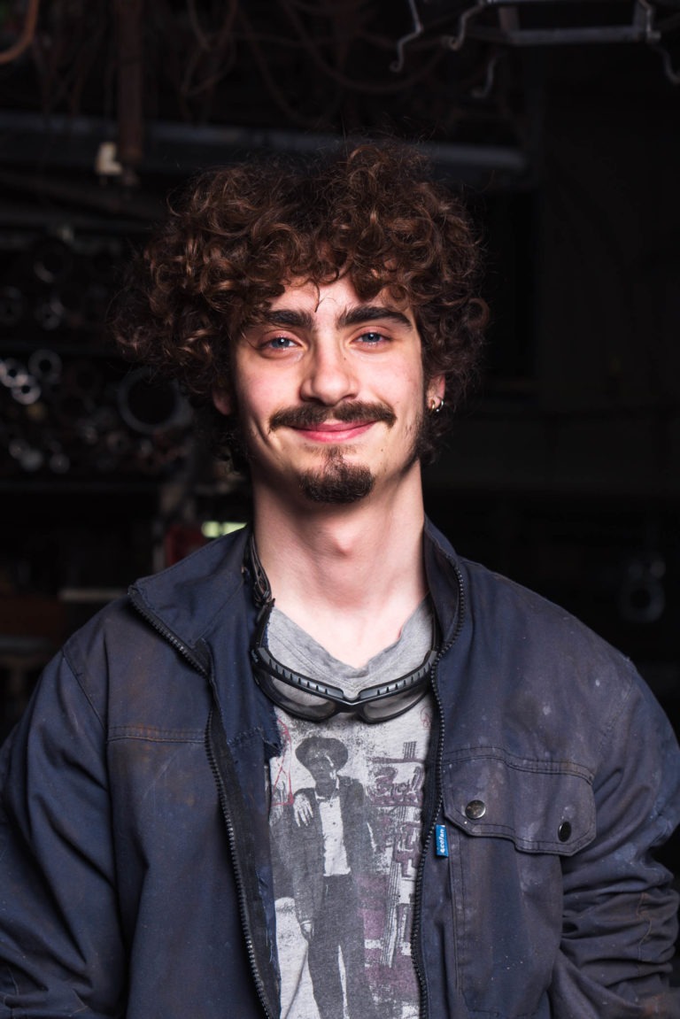 Un jeune homme aux cheveux bruns bouclés et au bouc sourit légèrement. Il porte une veste bleu foncé sur un t-shirt graphique représentant une silhouette d'homme. Autour de son cou pend une paire de lunettes de protection, faisant allusion à son travail à la Ferronnerie d'Art de Bordeaux. L'arrière-plan est faiblement éclairé, montrant des outils et des machines métalliques.