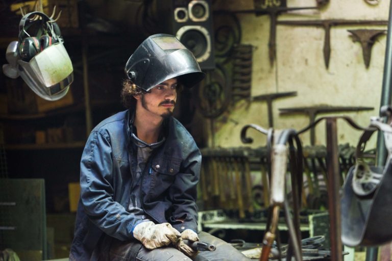 Un homme aux cheveux bouclés, portant un casque de soudeur sombre relevé et une veste en jean bleu, est assis dans un atelier encombré de la Ferronnerie d'Art de Bordeaux. Il tient une pièce de métal et porte des gants de protection. Divers outils et équipements sont visibles en arrière-plan, notamment un autre casque suspendu au-dessus de lui.