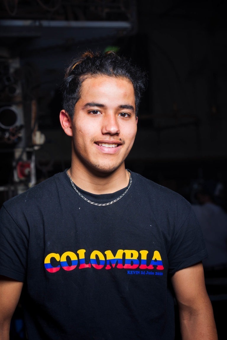 Un jeune homme aux cheveux noirs sourit à l'intérieur. Il porte un T-shirt noir avec le mot « COLOMBIA » en lettres colorées et audacieuses, aux couleurs du drapeau national (jaune, bleu et rouge). Le nom « KEVIN », la date « 24 juin 2023 » et un petit logo de la Ferronnerie d'Art Bordeaux sont imprimés en bas à droite du T-shirt