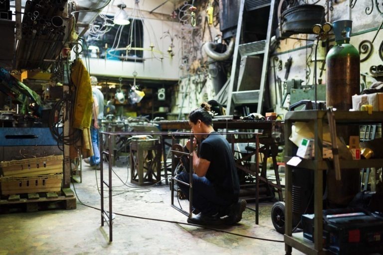 Une personne est agenouillée et soude du métal dans un atelier encombré de la Ferronnerie d'Art de Bordeaux. L'espace est rempli de divers outils, équipements et pièces métalliques. Des étincelles de soudure jaillissent près de la personne. L'arrière-plan comprend des étagères, des machines et un autre ouvrier. L'atmosphère est industrielle et animée.