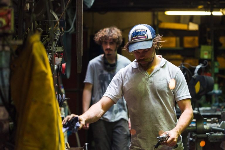 Deux hommes travaillent dans un atelier de la Ferronnerie d'Art de Bordeaux, faiblement éclairé, rempli d'outils et d'équipements. L'homme au premier plan, vêtu d'un polo blanc sale et d'une casquette de baseball, se concentre sur une tâche qu'il a entre les mains. À l'arrière-plan, légèrement flou, un homme aux cheveux bouclés et portant une chemise grise continue son travail.