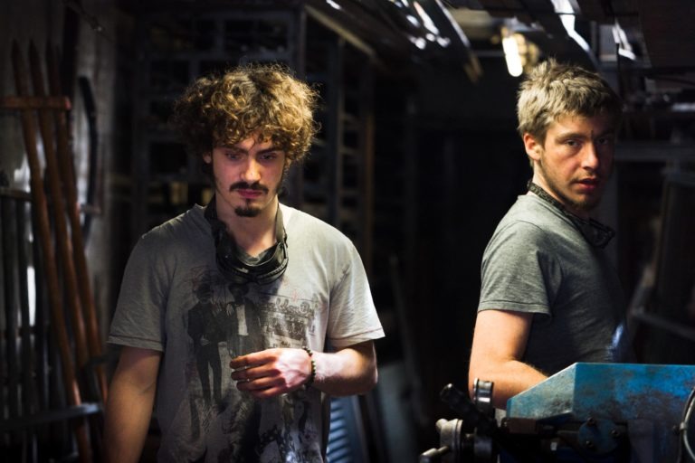 Deux hommes aux vêtements usés et aux expressions sérieuses se tiennent dans un atelier faiblement éclairé et encombré. L'homme de gauche a les cheveux bouclés, une barbe et porte un t-shirt gris avec un dessin, tenant un masque de soudure. L'homme de droite a les cheveux courts et un t-shirt gris, travaillant sur des machines bleues à la Ferronnerie d'Art de Bordeaux.