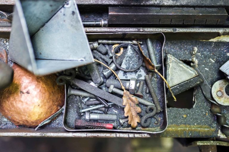 Un tiroir en métal avec divers objets, dont des vis, des boulons, une lime en métal, des triangles en métal, une sphère en cuivre et des feuilles de chêne tombées. La scène représente un environnement d'atelier de Ferronnerie d'Art Bordeaux en désordre avec des outils et des pièces éparpillés au hasard dans le tiroir.