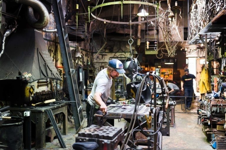 Un atelier rempli d'outils et d'équipements pour le travail des métaux. Au premier plan, une personne portant une chemise blanche et une casquette bleue travaille sur une pièce pour la Ferronnerie d'Art Bordeaux. L'atelier est encombré de pièces métalliques, d'étagères et de machines. À l'arrière-plan, on peut voir une autre personne travailler près de l'arrière de la boutique.