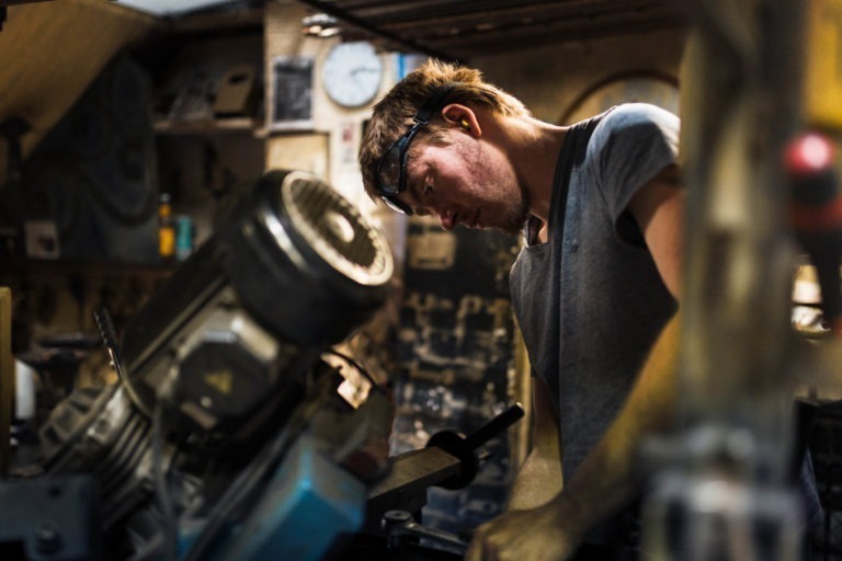 Un homme portant des lunettes de sécurité sur le front se penche en avant, concentré sur une tâche d'atelier à la Ferronnerie d'Art Bordeaux. Il porte un t-shirt gris et l'arrière-plan de l'atelier présente divers outils, équipements et machines industrielles. L'atmosphère tamisée crée une ambiance concentrée et industrieuse.