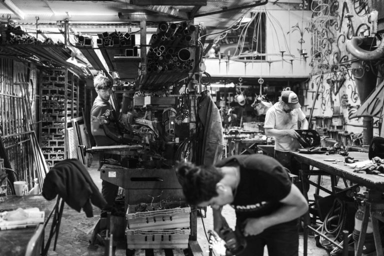 Image en noir et blanc de l'atelier de la Ferronnerie d'Art de Bordeaux. Trois personnes travaillent à différents postes, entourées d'outils et de pièces métalliques. Une personne au premier plan est penchée sur un établi, une autre utilise une machine et la troisième personne à droite est concentrée sur une tâche. L'espace est encombré et animé.