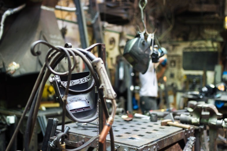 Un atelier encombré présente divers outils et équipements de travail du métal. Sur un établi en métal au premier plan, un masque de soudure et des fils sont bien en évidence. À l'arrière-plan, un artisan de la Ferronnerie d'Art Bordeaux en chemise blanche semble travailler, entouré de machines et de matériaux industriels.