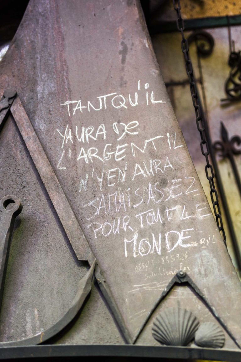 Une pièce en métal de la Ferronnerie d'Art Bordeaux porte le texte français écrit à la craie : « Tant qu'il y aura de l'argent, il n'y en aura jamais assez pour tout le monde. » Le métal présente des signes d'usure, avec une chaîne qui pend sur le côté droit et d'autres objets en métal vieilli en arrière-plan.