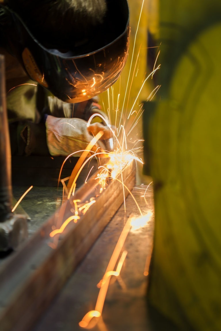 Un soudeur portant un équipement de protection, notamment un casque de soudage et des gants, est montré en train de souder du métal, créant des étincelles brillantes. L'image capture l'intensité de la Ferronnerie d'Art Bordeaux avec des étincelles vives et volantes sur un arrière-plan flou, soulignant la précision et l'énergie impliquées dans le soudage.