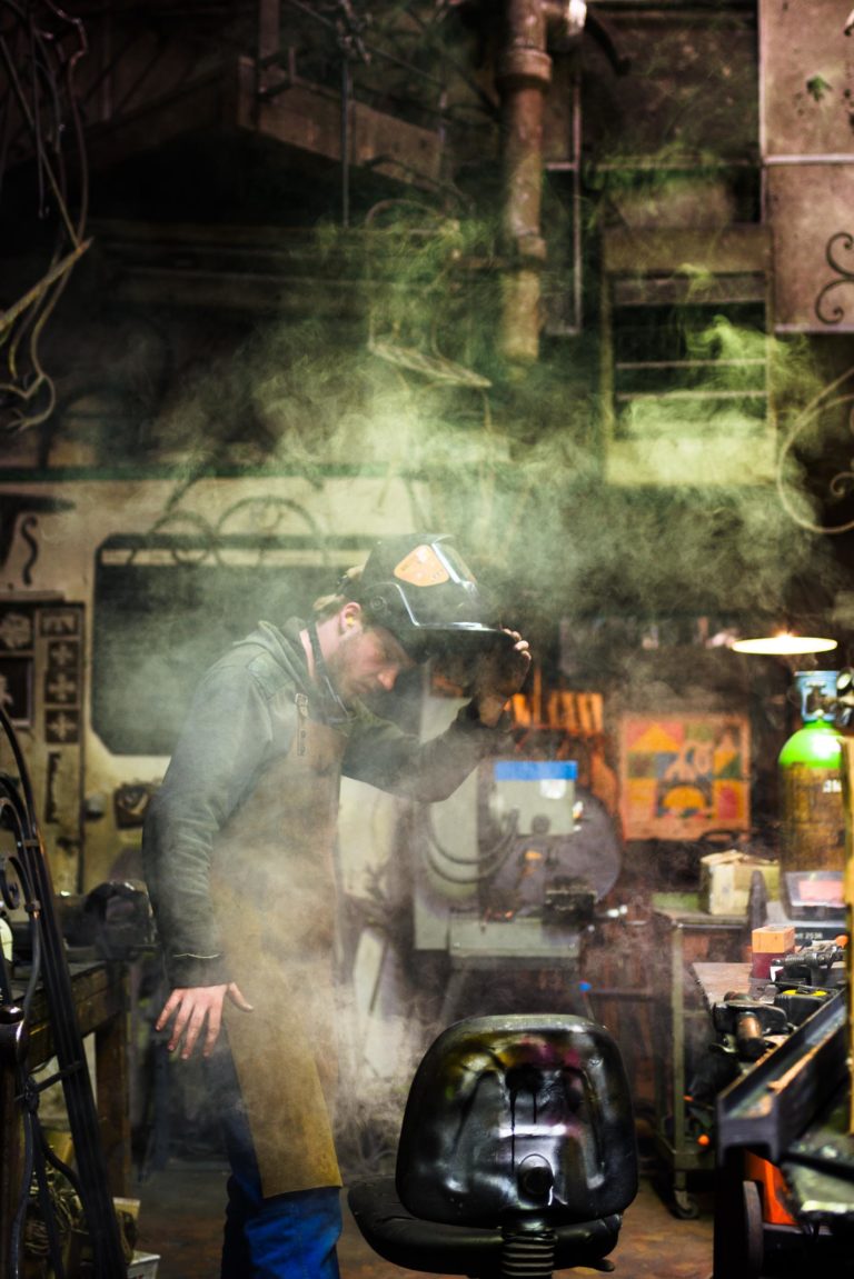 Dans un atelier, une personne, vêtue d'une veste et d'un tablier sombres, ajuste un casque de soudage. Une épaisse fumée ou vapeur remplit la zone. Divers outils, équipements et affiches colorées sont visibles en arrière-plan. L'atmosphère industrielle et brute de la Ferronnerie d'Art Bordeaux est illuminée par un éclairage d'ambiance.