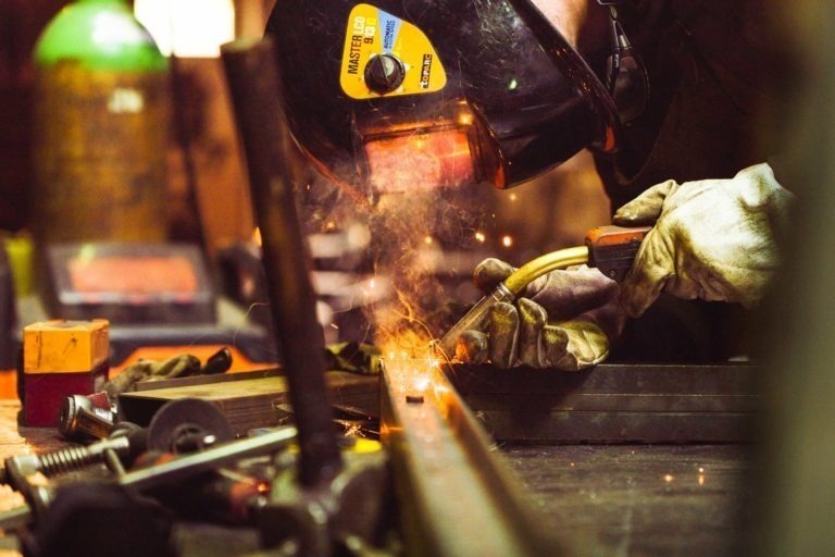 Un ouvrier portant un casque de soudeur et des gants de protection utilise un chalumeau sur une surface métallique à la Ferronnerie d'Art de Bordeaux, émettant des étincelles brillantes. Divers outils et équipements, dont des pinces et des pièces métalliques, sont éparpillés sur l'établi au premier plan. L'arrière-plan est légèrement flou, avec des éléments industriels.