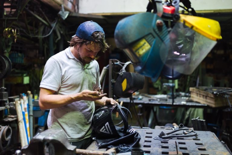 Dans un atelier, un homme examine une petite pièce dans sa main. Il porte un t-shirt blanc, une casquette bleue à l'envers et un casque de soudeur posé sur la table devant lui. L'espace, semblable à celui de la Ferronnerie d'Art de Bordeaux, est rempli d'outils et d'équipements, dont une grande perceuse à colonne et divers outils à main accrochés au mur.