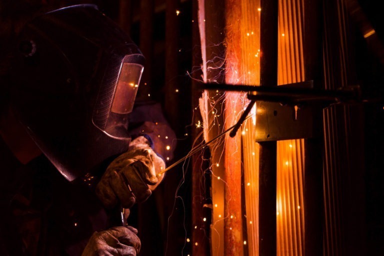 Un soudeur portant des gants de protection et un casque soude un joint métallique, produisant des étincelles brillantes et des panaches de fumée dans un atelier faiblement éclairé. La lueur orange du chalumeau illumine les structures métalliques environnantes et la silhouette de l'ouvrier, mettant en valeur le savoir-faire de la Ferronnerie d'Art Bordeaux.