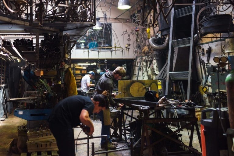 Un atelier animé de la Ferronnerie d'Art de Bordeaux avec trois personnes travaillant sur divers projets en métal. L'espace est rempli d'outils, de structures métalliques et de machines. Le personnage central soude, tandis qu'un autre à l'arrière-plan mesure quelque chose. L'environnement général est encombré et industrieux.