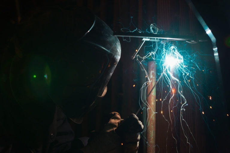 Une personne portant un casque de soudeur est en train de travailler à la Ferronnerie d'Art de Bordeaux. Des étincelles bleues et blanches illuminent la scène, mettant en valeur l'arc de soudage et créant un contraste saisissant avec l'environnement sombre. Le visage de l'individu n'est pas visible en raison de l'équipement de protection et de l'éclairage.