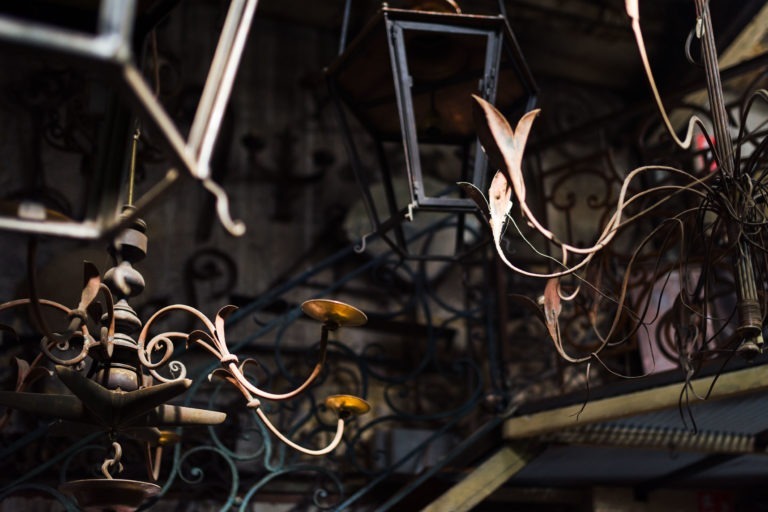 Un assortiment de lustres et de luminaires en métal anciens est suspendu à un haut plafond. En arrière-plan, on peut voir un escalier en fer forgé avec des rampes complexes de Ferronnerie d'Art Bordeaux. L'image est faiblement éclairée, soulignant l'atmosphère rustique et vintage du décor.