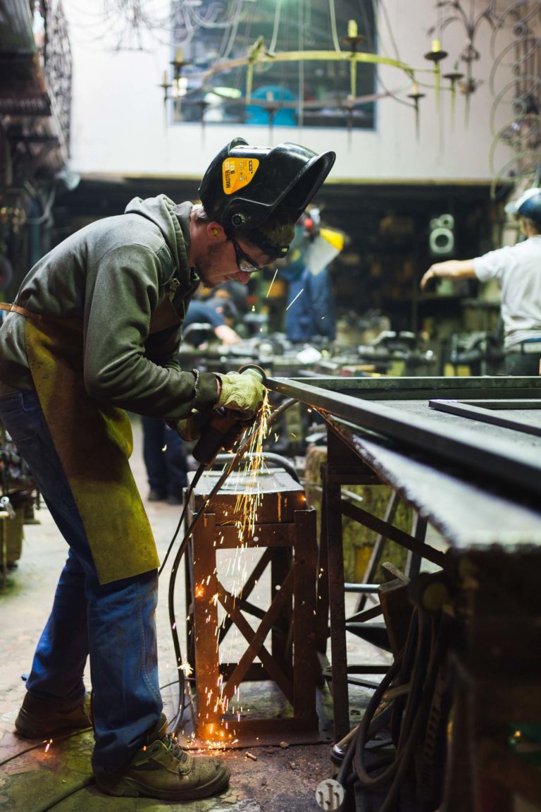 Une personne portant un casque de soudeur, des lunettes de sécurité et un équipement de protection utilise une meuleuse d'angle sur une pièce en métal à la Ferronnerie d'Art Bordeaux, créant des étincelles brillantes. L'atelier dispose de divers outils, équipements et d'autres travailleurs en arrière-plan. L'espace de travail est encombré de métal et de machines.