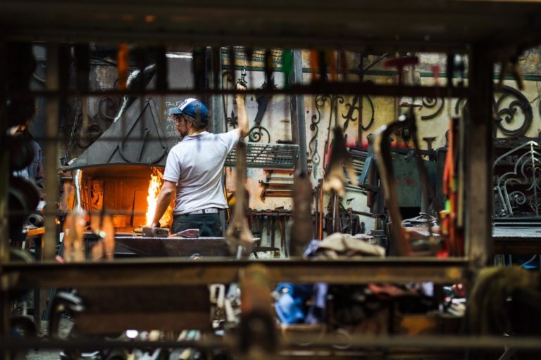 Une personne portant un casque bleu et un t-shirt blanc travaille dans une forge encombrée de la Ferronnerie d'Art de Bordeaux. Elle se tient devant un four ouvert avec une flamme orange vif, entourée de divers outils et objets en métal. La personne semble s'occuper du feu ou forger du métal, avec des cadres métalliques bloquant partiellement la vue.