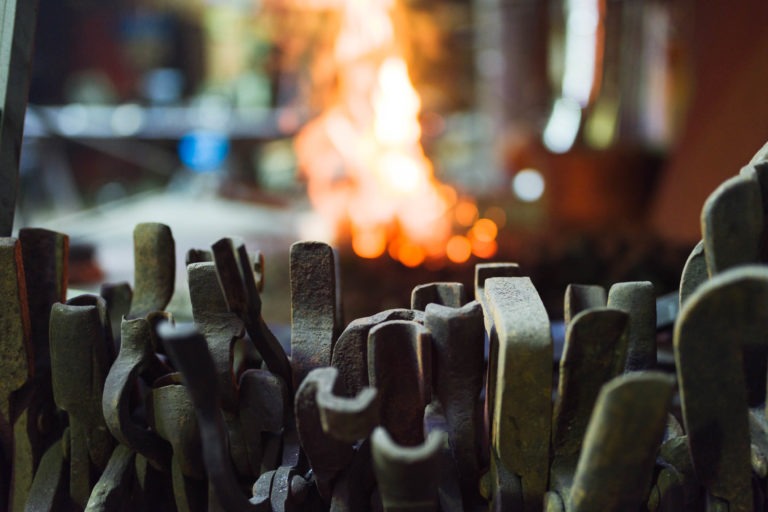 Au premier plan, une collection d'outils de forge robustes, avec une forge en feu en arrière-plan. Les diverses pinces et marteaux de la Ferronnerie d'Art Bordeaux sont légèrement usés par l'utilisation. Les flammes orange et jaune vibrantes contrastent fortement avec les outils en métal plus foncé.