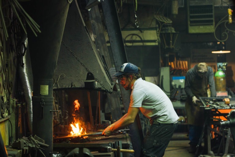 Un forgeron en t-shirt blanc et casquette travaille avec un outil à la forge, les flammes dansant dans l'âtre. L'atelier encombré de la Ferronnerie d'Art Bordeaux est parsemé d'outils et d'équipements, avec une autre personne en arrière-plan concentrée sur son propre poste.