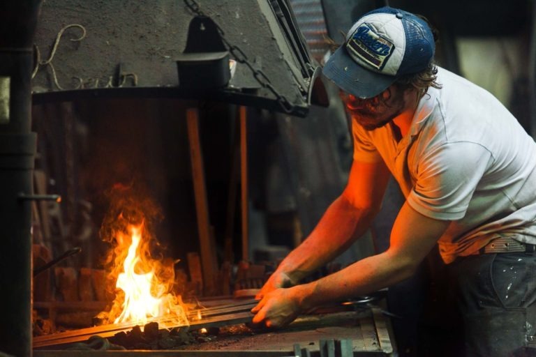 Un homme portant une casquette de camionneur bleue et blanche et un t-shirt blanc travaille sur une enclume dans une forge faiblement éclairée. Il tient une tige de métal dans le feu, ce qui fait danser des flammes orange vif. Ses manches sont retroussées alors qu'il se concentre sur sa tâche au milieu de divers outils, mettant en valeur le talent artistique de la Ferronnerie d'Art Bordeaux.