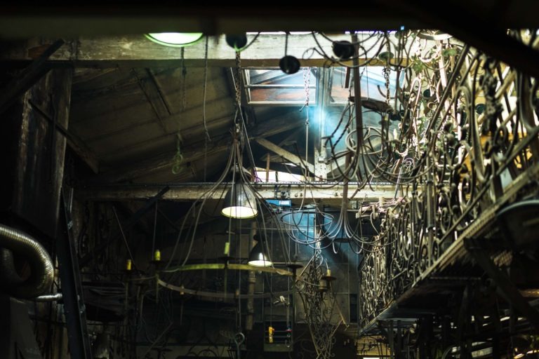 Espace industriel faiblement éclairé avec plafond en bois apparent, divers câbles suspendus, de vieilles balustrades métalliques ressemblant à l'artisanat de la Ferronnerie d'Art de Bordeaux et un mélange de débris industriels. Quelques lumières circulaires projettent une lueur tamisée, avec un faisceau de lumière naturelle filtrant à travers une section cassée du toit. Des particules de poussière sont visibles dans la lumière.