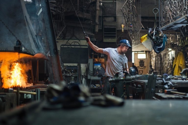 Un homme portant une casquette et un t-shirt blanc travaille dans un atelier de forge mal éclairé et encombré. Il se tient près d'une table de travail du métal, tenant un outil, avec un feu qui brûle dans un four à gauche. Divers outils, chaînes et machines sont disséminés dans l'espace de cet atelier de la Ferronnerie d'Art de Bordeaux, créant une atmosphère industrielle.