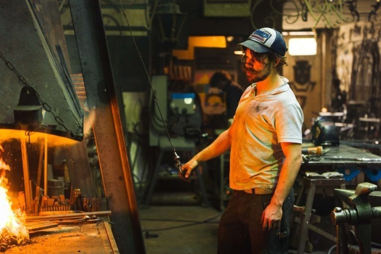 Un homme dans un atelier travaille avec une tige métallique tenue au-dessus d'une flamme nue. Il porte une casquette de sécurité, des lunettes de protection et une chemise blanche. L'arrière-plan est rempli de divers outils, équipements et d'un établi, incarnant l'esprit industriel de la Ferronnerie d'Art Bordeaux. Un casque de soudeur est posé sur l'établi.