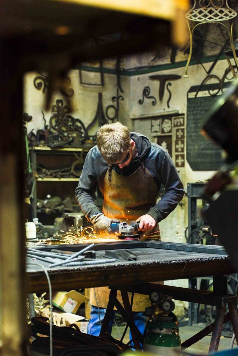 Une personne portant un sweat à capuche et des lunettes de sécurité utilise une meuleuse, créant des étincelles, dans l'atelier de la Ferronnerie d'Art Bordeaux rempli d'outils de travail du métal et de ferronnerie décorative sur les murs. La personne est concentrée sur son travail à une table encombrée de pièces et d'équipements métalliques, éclairée par l'éclairage de l'atelier.