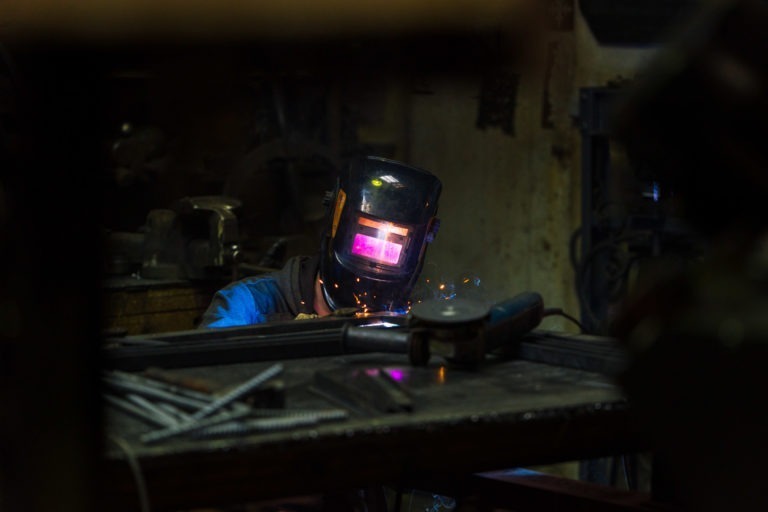 Un soudeur portant un casque sombre et un équipement de protection travaille sur du métal dans un atelier de la Ferronnerie d'Art de Bordeaux faiblement éclairé. La visière de son casque brille sous l'effet des étincelles de soudure. Divers outils et tiges métalliques sont éparpillés sur un établi au premier plan, tandis que l'arrière-plan est encombré d'équipements industriels.