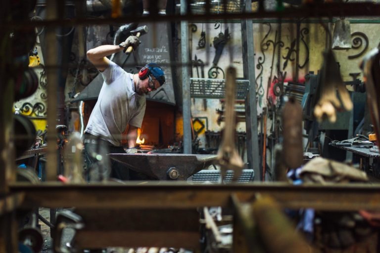 Un forgeron, coiffé d'un casque de protection et d'un cache-oreilles, travaille sur une pièce de métal posée sur une enclume dans un atelier encombré et faiblement éclairé. Il tient un marteau et travaille près d'une forge rougeoyante. Divers outils et pièces métalliques de la Ferronnerie d'Art Bordeaux sont accrochés aux murs et éparpillés un peu partout.