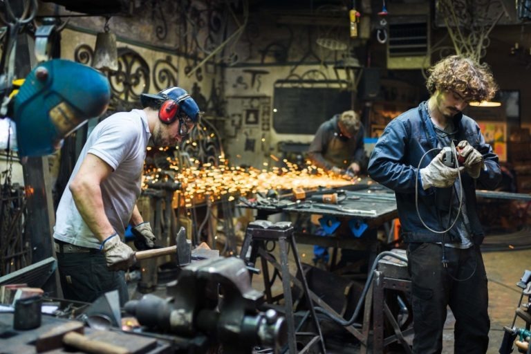 Deux ouvriers travaillent dans un atelier encombré. L'ouvrier de gauche, portant des protections auditives et une chemise blanche, utilise une meuleuse d'angle, produisant des étincelles. L'ouvrier de droite, aux cheveux bouclés et portant une veste sombre, travaille avec un outil à main. Ferronnerie d'Art Bordeaux remplit l'espace de travail d'outils et d'équipements.