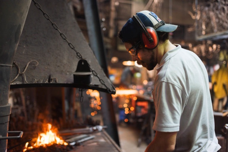 Un forgeron vêtu d'une chemise blanche et d'un équipement de protection, notamment des cache-oreilles, des lunettes de sécurité et une casquette bleue estampillée « Ferronnerie d'Art Bordeaux », travaille dans une forge faiblement éclairée. Il se concentre sur un feu à son poste de travail, avec divers outils et équipements visibles en arrière-plan, éclairés par la lumière ambiante.