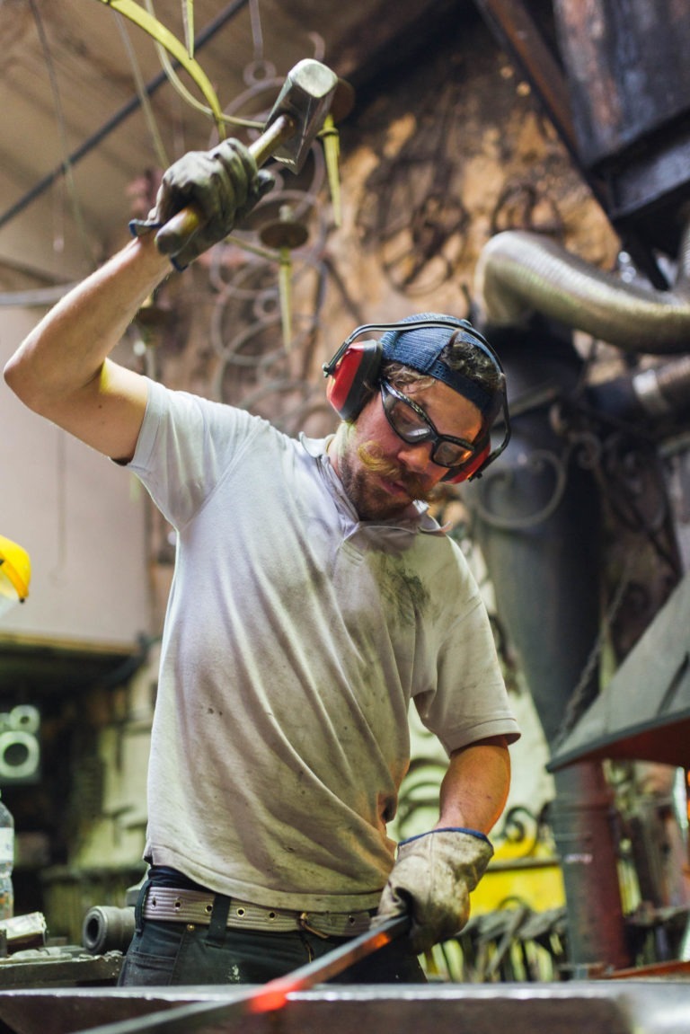 Un forgeron de la Ferronnerie d'Art de Bordeaux, portant des lunettes de protection, des cache-oreilles et des gants, frappe une pièce de métal brûlante avec un marteau. L'arrière-plan de l'atelier présente divers outils et équipements. L'expression concentrée du forgeron et son effort physique soulignent l'intensité du processus de travail du métal.