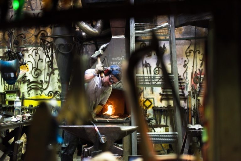 Un forgeron portant un équipement de protection, notamment des gants et un bandana, travaille dans un atelier encombré rempli d'outils et de décorations complexes de la Ferronnerie d'Art Bordeaux. Il manie un marteau près d'une enclume avec une forge rougeoyante en arrière-plan, soulignant la chaleur intense et le savoir-faire impliqué dans le travail du métal.