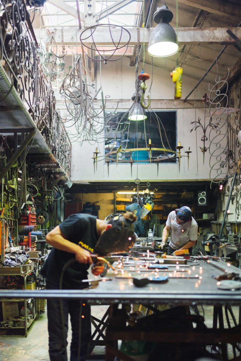 Deux personnes travaillent sur de grandes structures métalliques dans un atelier rempli d'outils et de pièces de métal complexes. Ils portent des casques de sécurité et utilisent du matériel de soudage, créant des étincelles. La pièce est encombrée de pièces métalliques, de balustrades et d'objets suspendus sous un éclairage industriel à la Ferronnerie d'Art Bordeaux.