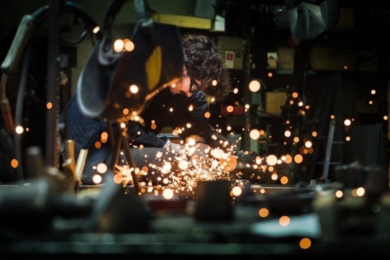 Dans un atelier faiblement éclairé, une personne portant un équipement de sécurité, notamment des lunettes de protection, utilise un outil électrique pour meuler du métal, créant une pluie d'étincelles brillantes. Les environs de la Ferronnerie d'Art Bordeaux sont remplis d'outils et d'équipements divers, les étincelles ajoutant du contraste et du mouvement dynamique à la scène.