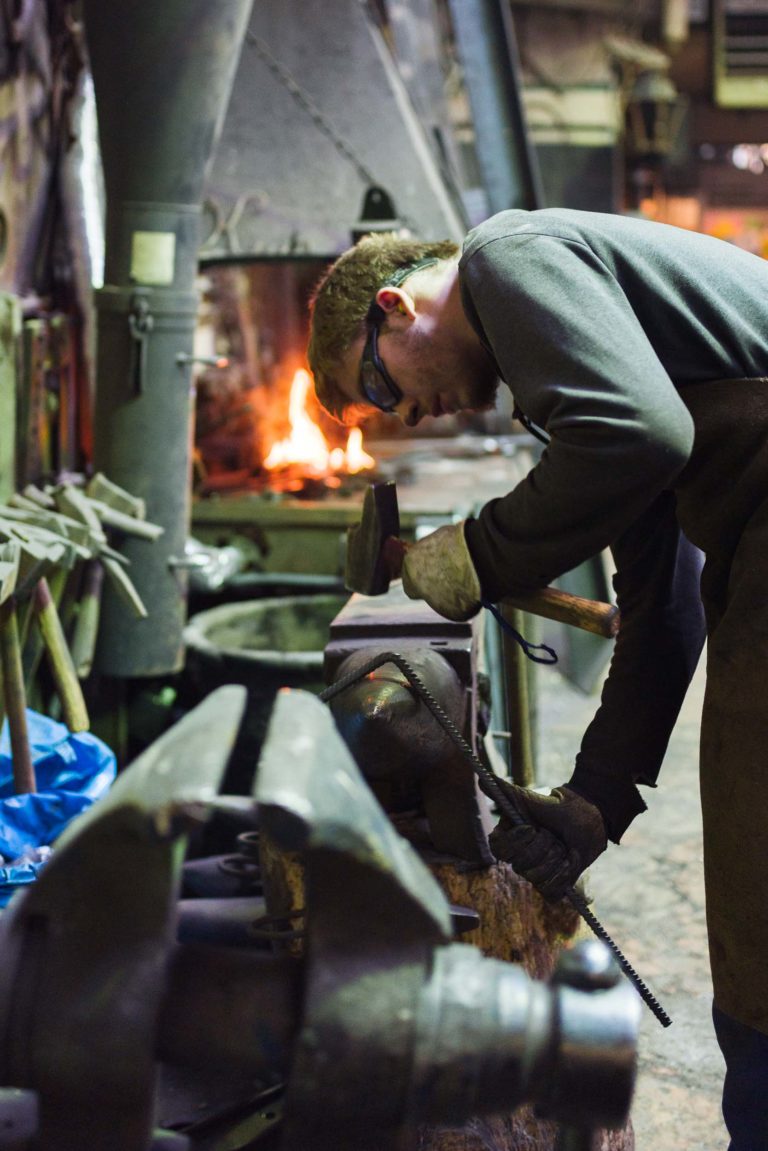 Dans l'atelier animé de la Ferronnerie d'Art Bordeaux, un forgeron portant des lunettes de sécurité et une chemise sombre martèle une pièce de métal sur une enclume. Divers outils sont visibles au premier plan, tandis que les flammes orange vif d'une forge illuminent les étagères encombrées et les équipements qui tapissent les murs.