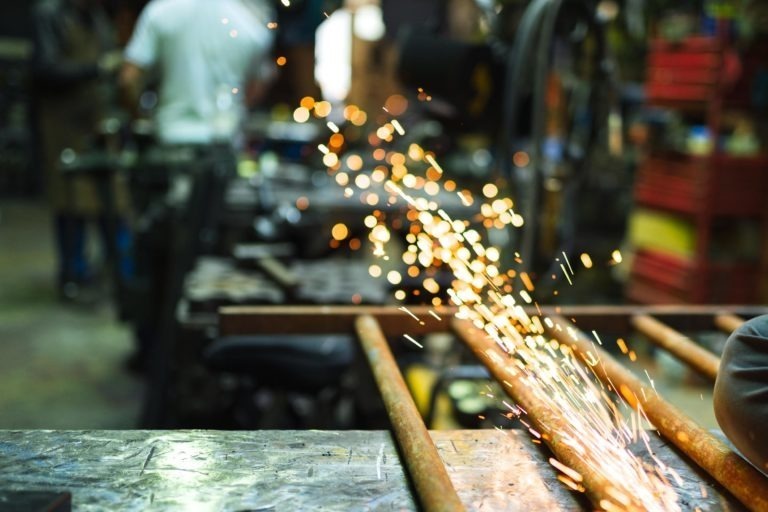 Une scène d'atelier avec une personne manipulant une meuleuse sur une tige métallique, créant une pluie d'étincelles orange vif. L'arrière-plan présente divers outils, équipements et étagères remplies de matériaux, le tout légèrement flou, soulignant l'environnement actif et dynamique de la Ferronnerie d'Art Bordeaux.