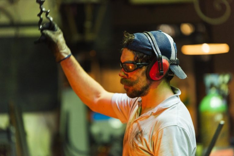 Un homme portant un équipement de sécurité, notamment des protections auditives, une casquette et des lunettes de protection, travaille dans une Ferronnerie d'Art industrielle de Bordeaux. Il porte une moustache et sa chemise est tachée, ce qui indique un travail manuel ou un atelier. Il tient un outil dans sa main gantée, avec un équipement industriel flou en arrière-plan.