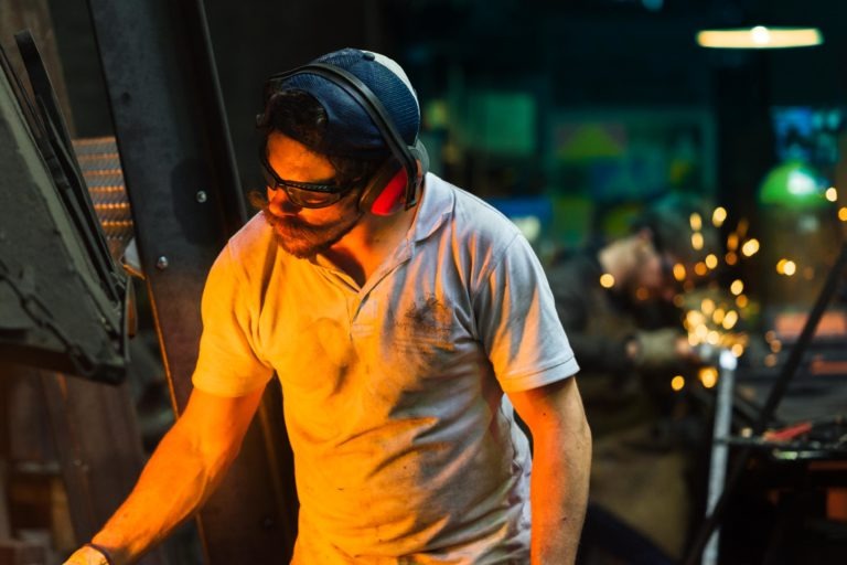 Une personne portant une casquette, des lunettes de sécurité et des protections auditives travaille dans un atelier faiblement éclairé. Vêtue d'une chemise blanche sale, elle semble concentrée. En arrière-plan, un collègue de la Ferronnerie d'Art Bordeaux travaille avec des outils, émettant des étincelles visibles. Tous deux sont entourés d'équipements et de machines industrielles.