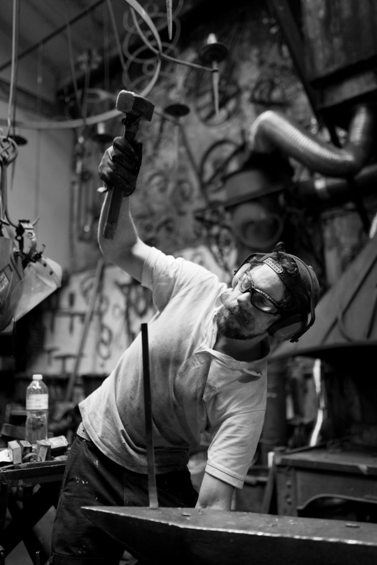 Image en noir et blanc d'un forgeron au travail dans un atelier. Il porte des gants de protection, des lunettes de sécurité et un casque de soudeur relevé. Il tient un marteau levé au-dessus d'une pièce de métal sur une enclume. L'arrière-plan présente divers outils, des machines et une bouteille étiquetée "Ferronnerie d'Art Bordeaux".