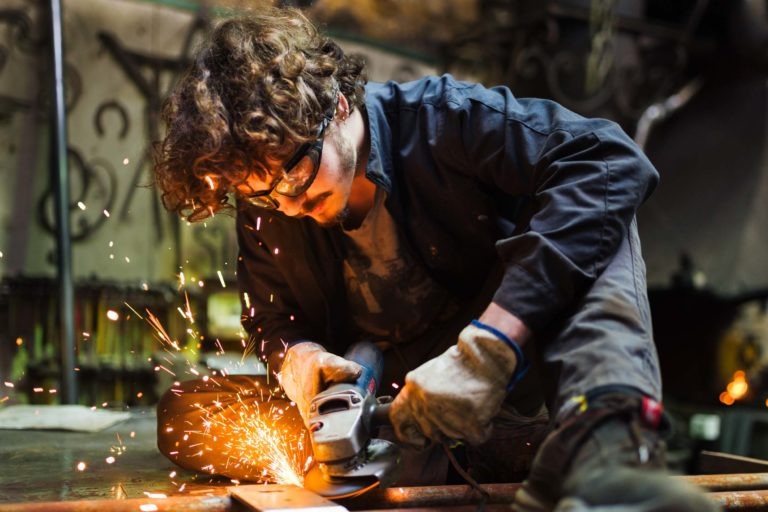 Une personne aux cheveux bouclés et aux lunettes de sécurité utilise une meuleuse d'angle, produisant des étincelles. Elle porte des gants, une veste sombre et est concentrée sur son travail, à genoux sur le sol dans un atelier rempli d'outils et d'équipements de la Ferronnerie d'Art Bordeaux.