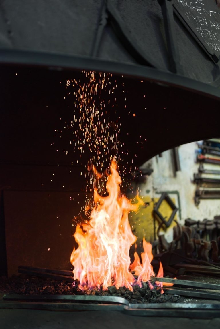 Une forge aux flammes orange et jaune vif s'intensifie au centre, produisant une cascade d'étincelles incandescentes. L'arrière-plan présente des outils et du matériel accrochés sur un mur légèrement flou, indiquant le cadre artisanal de la Ferronnerie d'Art Bordeaux.