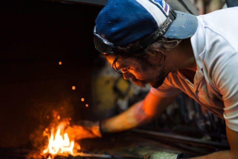 Une personne portant une chemise blanche, une casquette bleue et blanche et des lunettes de sécurité s'occupe d'un petit feu qui fait des étincelles dans un atelier de la Ferronnerie d'Art de Bordeaux faiblement éclairé. Tendant les mains gantées vers le feu, l'individu se concentre intensément sur son travail.