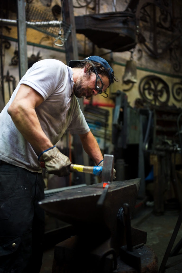 Un forgeron est concentré sur le travail du métal incandescent qu'il martèle sur une enclume. Il porte des gants de protection, une chemise blanche à manches courtes et une casquette bleue portée à l'envers. Divers outils et équipements remplissent l'atelier de la Ferronnerie d'Art de Bordeaux, créant une atmosphère rustique et industrieuse.