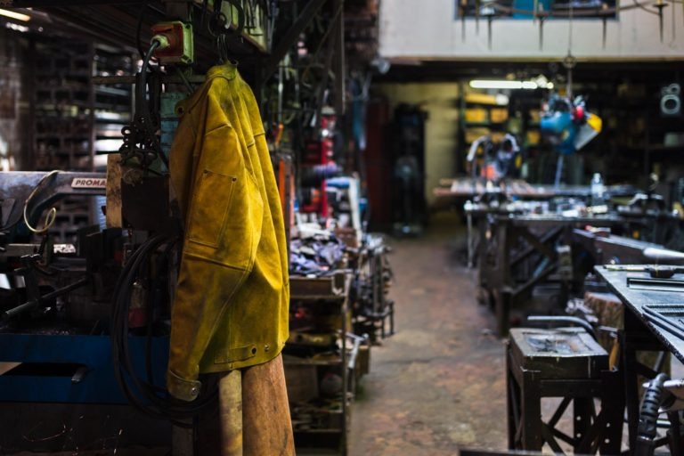 Une veste de travail jaune usée est suspendue à un crochet dans un atelier faiblement éclairé et encombré. L'espace, appartenant à la Ferronnerie d'Art Bordeaux, est rempli d'équipements de travail du métal et d'outils divers, avec des établis et des machines disséminés un peu partout. La scène suggère un espace de travail actif, peut-être industriel.