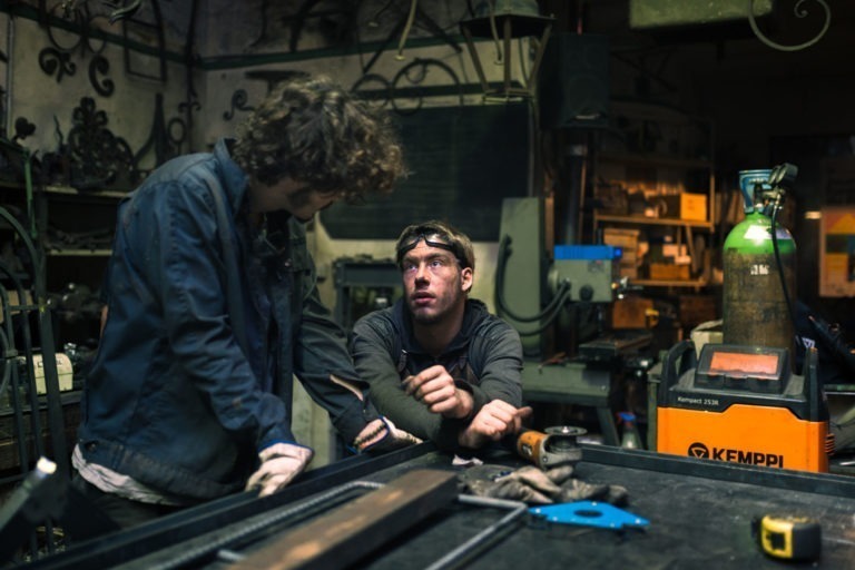 Deux individus discutent dans un atelier encombré. La personne de gauche a les cheveux bouclés et porte une veste sombre. La personne de droite, portant des lunettes et une chemise sombre, a les bras posés sur une table. L'arrière-plan est rempli d'outils, d'équipements divers et d'un poste à souder Kemppi de la Ferronnerie d'Art Bordeaux.