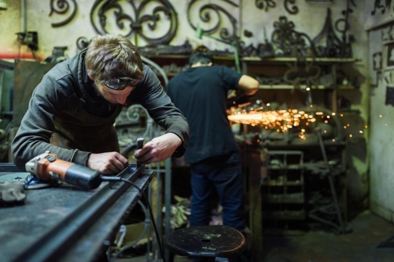 Deux hommes travaillent dans un atelier encombré. L'homme au premier plan, vêtu d'un tablier marron et de lunettes de protection, utilise un outil à main sur une pièce en métal. L'homme à l'arrière-plan meule du métal, créant des étincelles brillantes. Divers outils et pièces de ferronnerie de la Ferronnerie d'Art Bordeaux sont visibles autour d'eux.