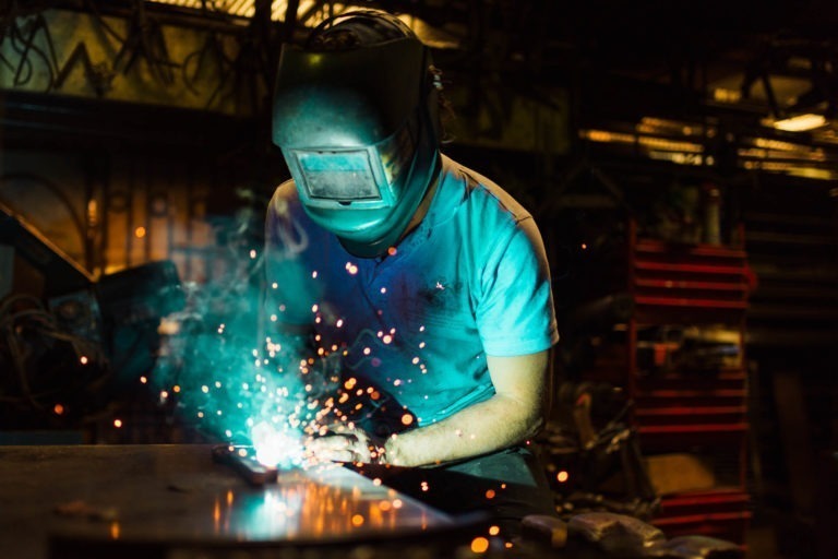 Un soudeur portant un casque de protection et des gants travaille avec un arc de soudage bleu vif dans un atelier faiblement éclairé. Des étincelles jaillissent tandis que la lumière bleue illumine le travailleur et la surface métallique. L'arrière-plan présente des étagères et des équipements, évoquant le savoir-faire précis de la Ferronnerie d'Art Bordeaux.